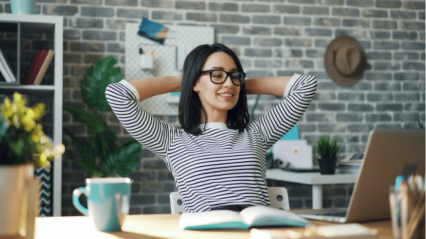 Femme souriante s'étirant les bras derrière la tête devant son ordinateur, illustrant le bien-être au travail.