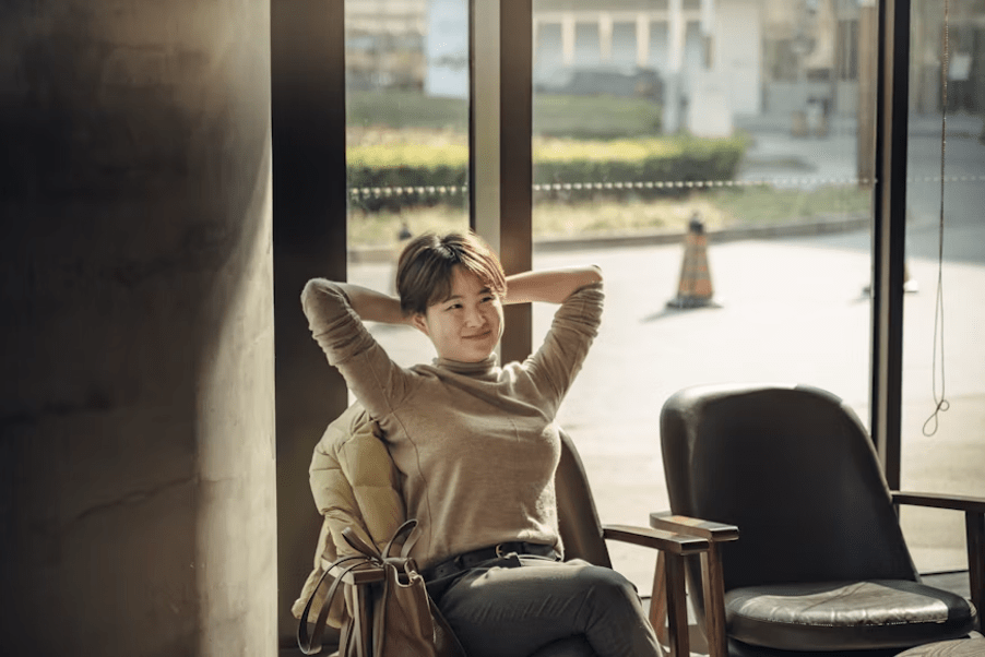 Femme souriante prenant une pause relaxante loin de son écran, assise près d'une grande fenêtre.